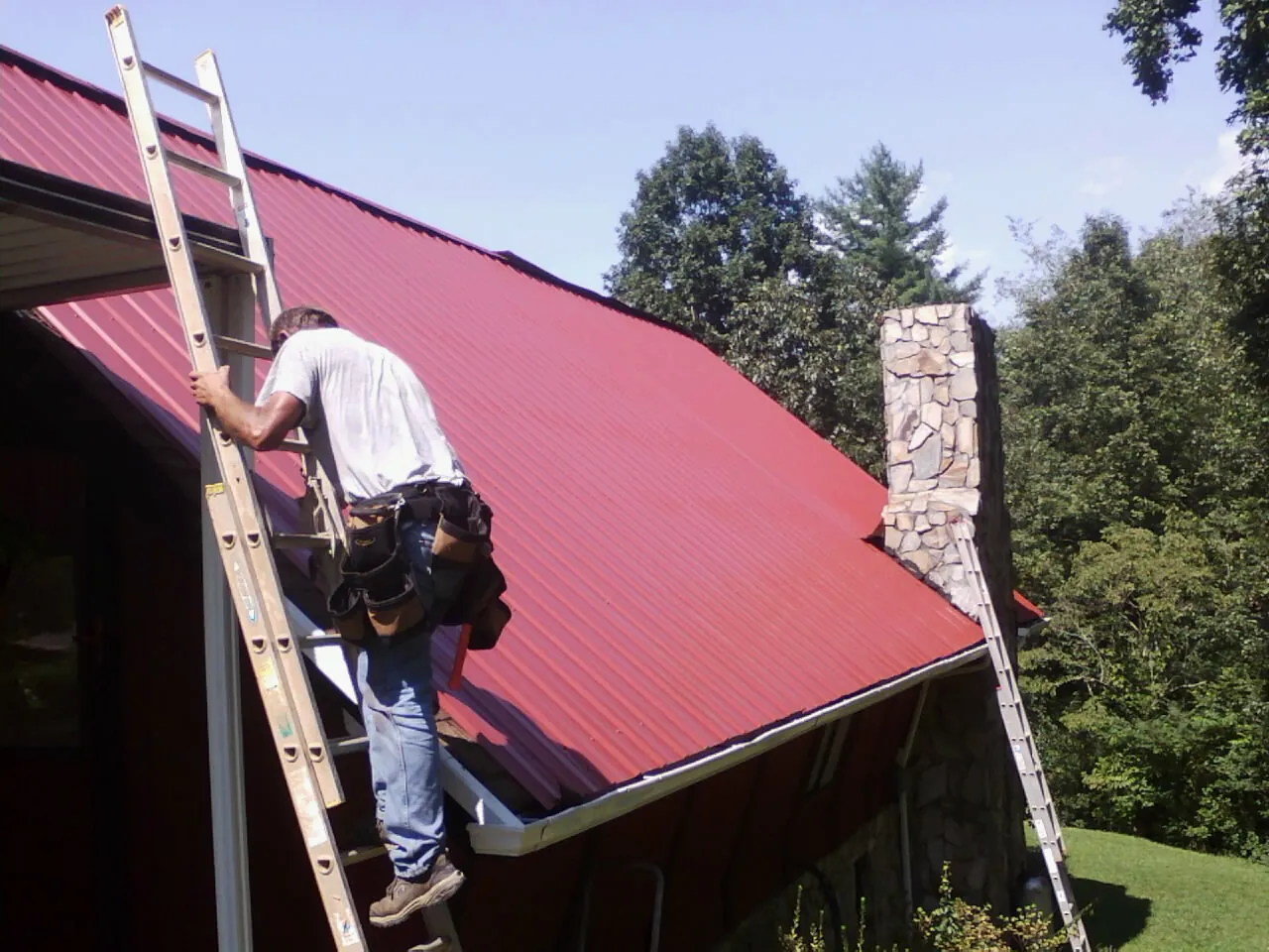 Roofer on a ladder beside a red metal roof and stone chimney — Soffit & Fascia Repair by Roofing Brothers in Hedgesville